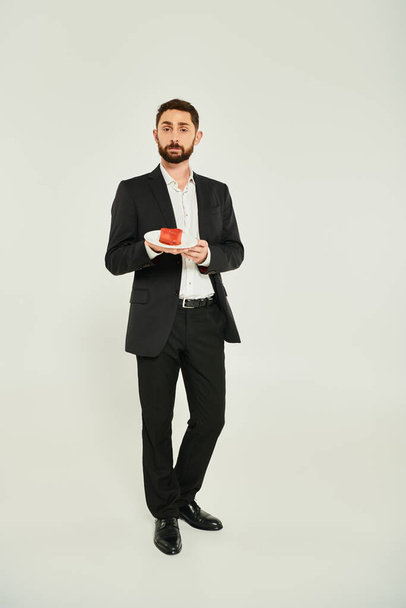 plná délka ostře oblečeného muže držícího talíř s dort ve tvaru srdce na šedé, st valentines koncept - Fotografie, Obrázek