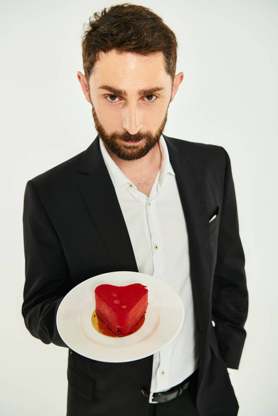 bearded elegant man showing plate with red heart-shaped cake on grey, st valentines day treat - Photo, Image