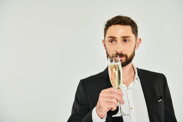 homem barbudo elegante em terno preto bebendo champanhe e olhando para longe no dia dos namorados cinza, st - Foto, Imagem