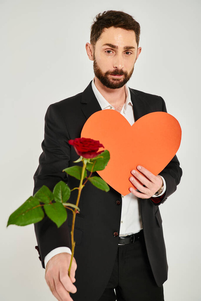 stylowy brodaty mężczyzna z czerwoną różą i pomarańczowym papierowym sercem patrząc na aparat fotograficzny na szarym, st walentynki - Zdjęcie, obraz