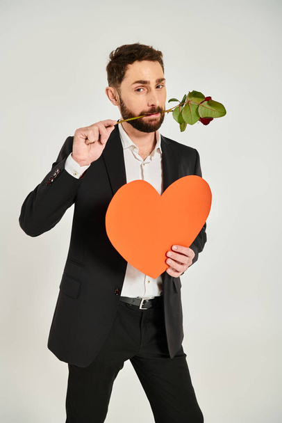 γενειοφόρος κομψός άνθρωπος με πορτοκαλί καρδιά χαρτί και κόκκινο τριαντάφυλλο στα δόντια σε γκρι, st Valentines έννοια - Φωτογραφία, εικόνα