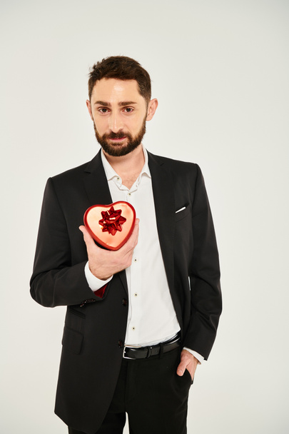 hombre barbudo positivo y elegante mostrando caja de regalo en forma de corazón con arco en gris, día de San Valentín - Foto, Imagen