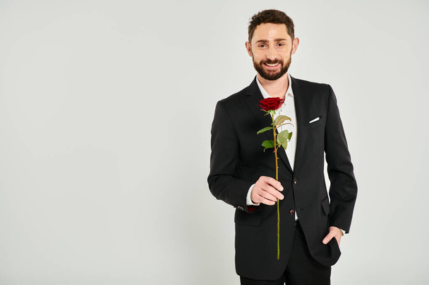 vidám szakállas férfi fekete formális viselet kezében vörös rózsa és a kamera szürke háttér - Fotó, kép