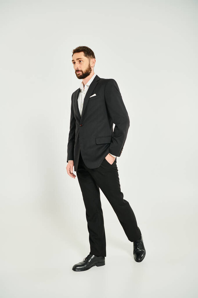 in voller Länge wohlhabender Geschäftsmann im schwarzen eleganten Anzug posiert mit der Hand in der Tasche auf grau - Foto, Bild