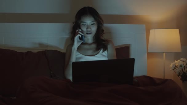 Średnie ujęcie młodej kobiety pracującej na własny rachunek dzwoniącej do kolegi i pytającej o szczegóły nowego projektu siedząc do późna w nocy przed laptopem w łóżku - Materiał filmowy, wideo