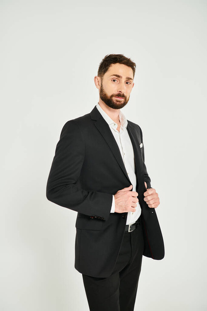 hombre de negocios carismático vestido agudo en ropa formal negro mirando a la cámara en el fondo gris - Foto, imagen