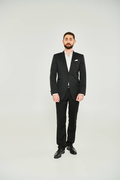 Vorderseite des bärtigen Geschäftsmannes im schwarzen eleganten Anzug, der vor grauem Hintergrund steht, in voller Länge - Foto, Bild