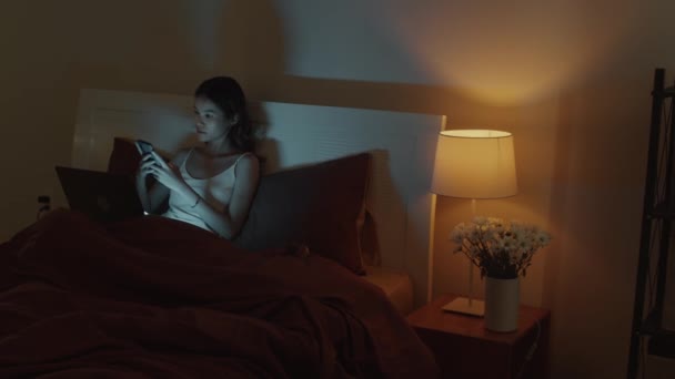 Genç Asyalı bir kadının dizüstü bilgisayarla çalışırken cep telefonuna baktığı orta boy bir fotoğraf. Geceleri yatakta oturuyor, uzayı kopyalıyor. - Video, Çekim