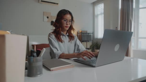 Medium shot van pensive vrouwelijke student doen cursus werk op laptop zitten aan het bureau in gezellige flat - Video
