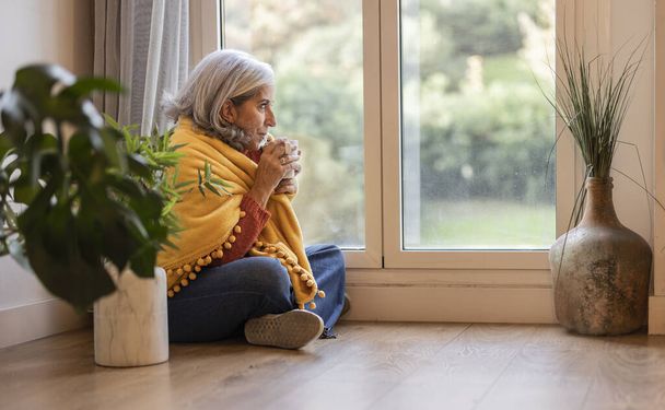 starsza kobieta siedzi na podłodze swojego mieszkania pijąc gorący napój przy oknie przykrytym - Zdjęcie, obraz