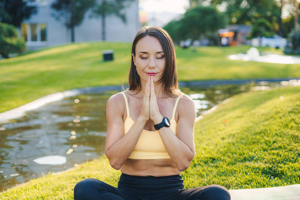 Portret dojrzałej brunetki uprawiającej jogę na świeżym powietrzu w zielonym parku i trzymającej się za ręce dla równowagi w pozycji modlitewnej. Ćwiczenia jogi i modlitwy lotosu - Zdjęcie, obraz