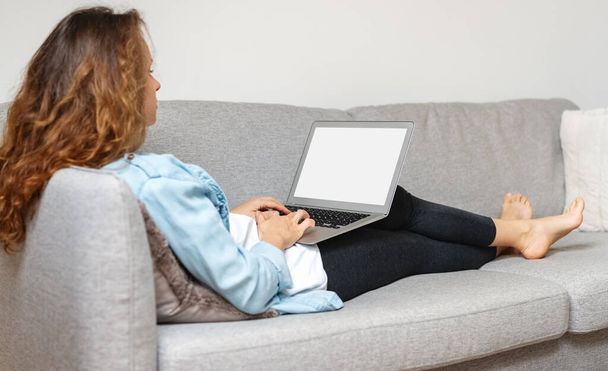 Vista trasera de la mujer sentada en el sofá y trabajando en el ordenador portátil. Pantalla en blanco del ordenador portátil. - Foto, imagen