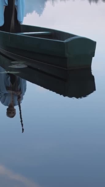 Vídeo vertical. El reflejo en el agua de un barco de madera con un hombre apoyado en un remo y flotando entre el agua con nenúfares y niebla. Concepto de transporte de almas después de la muerte. - Metraje, vídeo