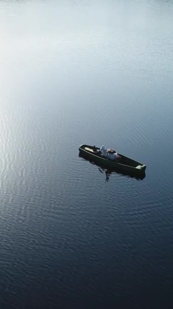 Pystysuora video. Mies soittaa kitaraa makatessaan puuveneessä keskellä villiä järveä veden pinnalla auringon säteiden heijastuessa aaltoihin. Antenninäköala. - Materiaali, video