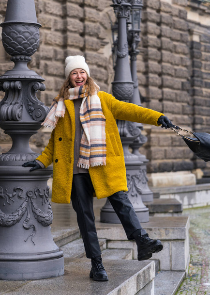 Stylish elegant woman in a yellow coat enjoys new places. Laughs merrily, holds the pole with his hand. Happy traveler woman looking at tourist attractions of Dresden city, Germany  - Photo, Image