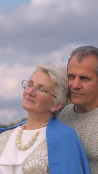 Vídeo vertical. Un hombre en edad de jubilación sostiene a una anciana con gafas en sus brazos. Se frota la cabeza en la cara de sus maridos. Miran a la distancia. Concepto de vida feliz. - Metraje, vídeo