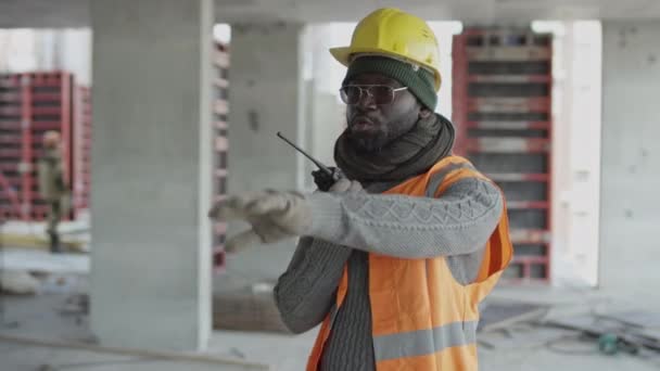 Keskikokoinen laukaus Musta mies rakennustyömaan esimies yllään kypärä ja turvaliivi tehdä käsi eleitä ja antaa ohjeet työmiesten osaksi radiopuhelin - Materiaali, video