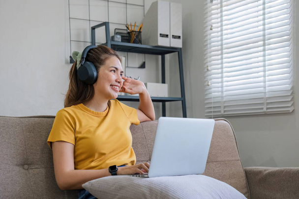 Aasialainen nainen istuu sohvalla olohuoneessa kannettavan tietokoneen avulla ja kuuntelee musiikkia kuulokkeilla.. - Valokuva, kuva