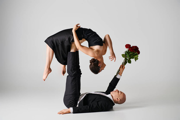 flessibile donna in abito nero bilanciamento sui piedi del partner di danza che tiene rose rosse su grigio - Foto, immagini