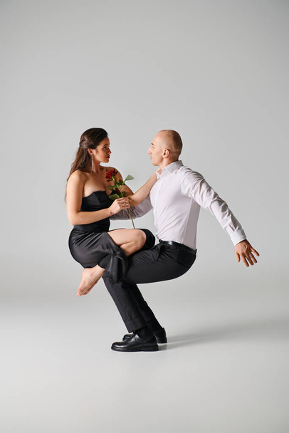 brunette woman in black dress holding red rose and balancing on laps of male dancer on grey backdrop - Photo, Image
