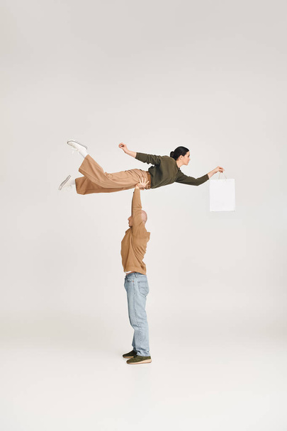 uomo forte in abbigliamento casual sollevamento donna con shopping bag, bilanciamento in una posa giocosa in studio - Foto, immagini