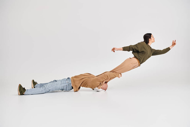 акробатка в повседневной одежде балансирует в воздухе при поддержке своего партнера в серой студии - Фото, изображение
