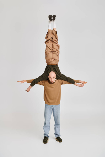 silny mężczyzna w luźnym ubraniu wspierające ciało partnera akrobaty podczas próby w studio na szarości - Zdjęcie, obraz