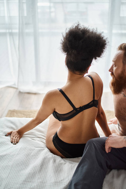 Многорасовая пара отдыхает на кровати в уютной комнате, кудрявая черная женщина в нижнем белье и бородатый мужчина - Фото, изображение