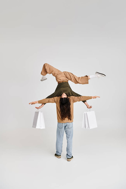 mujer acrobática sosteniendo compras y equilibrando al revés con el apoyo de la pareja en gris - Foto, imagen