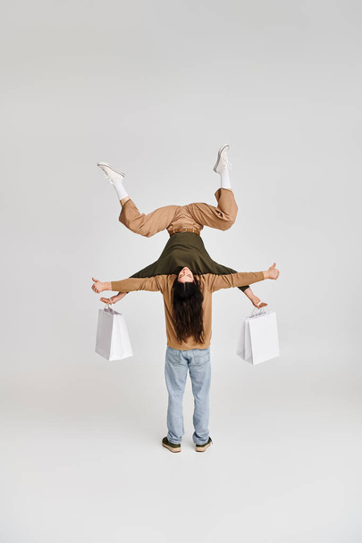 vrouw houdt boodschappentassen vast en balanceert ondersteboven met steun van acrobatische partner in studio - Foto, afbeelding