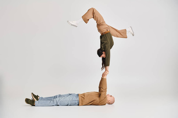 acrobatische uitvoering van artistiek paar, vrouw in casual kleding balanceren op handen van de man op grijs - Foto, afbeelding