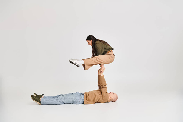 acrobatic performance of young couple, woman in casual clothes balancing on hands of man on grey - Photo, Image