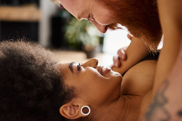 Χαρούμενη Αφροαμερικάνα που κοιτάζει τον γκόμενο με τα τατουάζ με τα γένια στην κρεβατοκάμαρα, προσωπικές στιγμές - Φωτογραφία, εικόνα