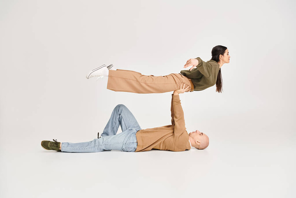 homem forte em casual desgaste deitado no chão e levantando mulher morena no estúdio, casal de acrobatas - Foto, Imagem