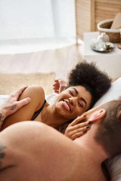 felice donna afroamericana sorridente mentre sdraiata sul letto insieme al suo fidanzato tatuato - Foto, immagini