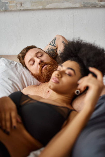 bearded man with tattoo looking at his african american girlfriend in lingerie lying next to him - Photo, Image