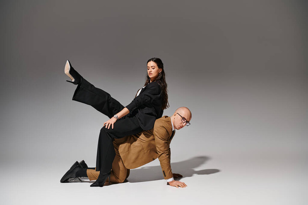 Zakelijk paar in stijlvolle pakken in creatieve acrobatische pose, vrouw zitten op de rug van de man in de studio - Foto, afbeelding