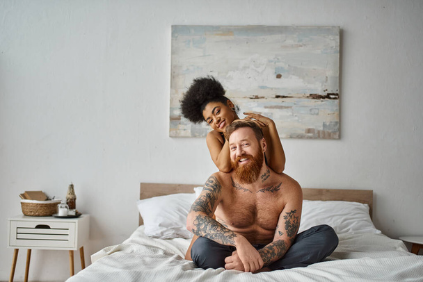 feliz pareja diversa, mujer afroamericana abrazando a su hombre tatuado en una cama en una habitación luminosa - Foto, Imagen
