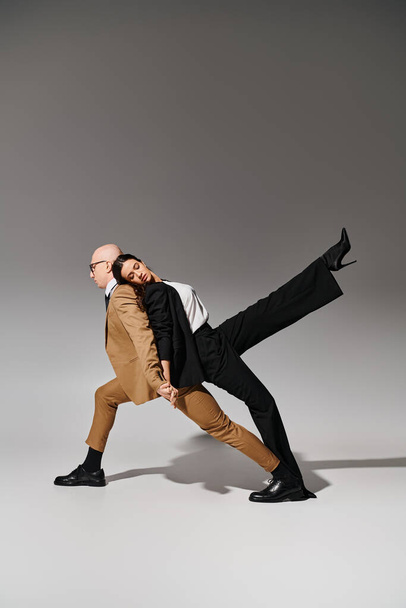 partners in coordinated dance move, woman in suit leaning on back of man in business attire on grey - Photo, Image