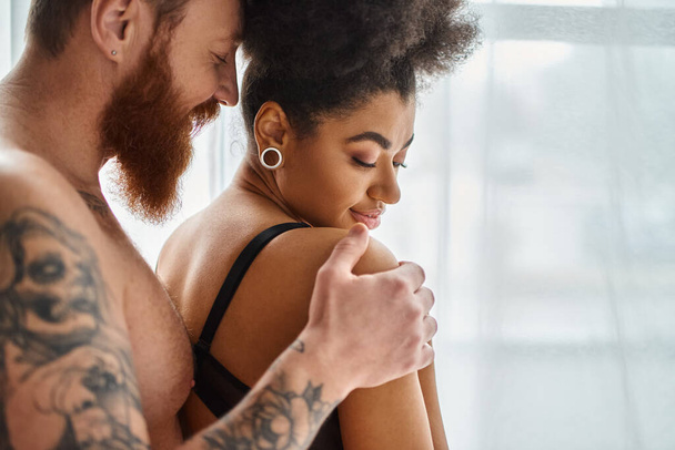 happy and shirtless man with beard touching shoulder of african american girlfriend in lingerie - Photo, Image
