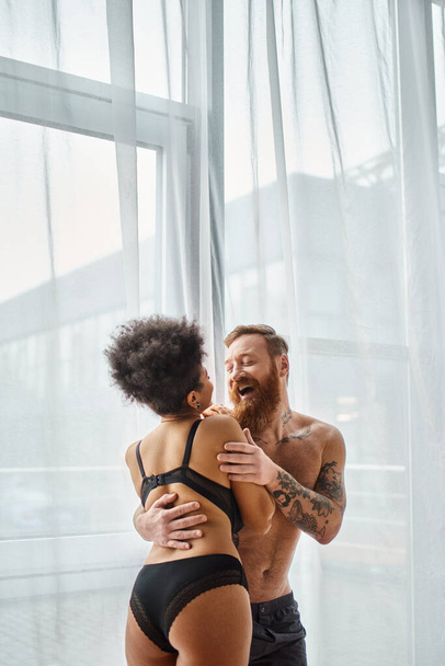 excited and tattooed man without shirt hugging his happy african american girlfriend near curtain - Photo, Image