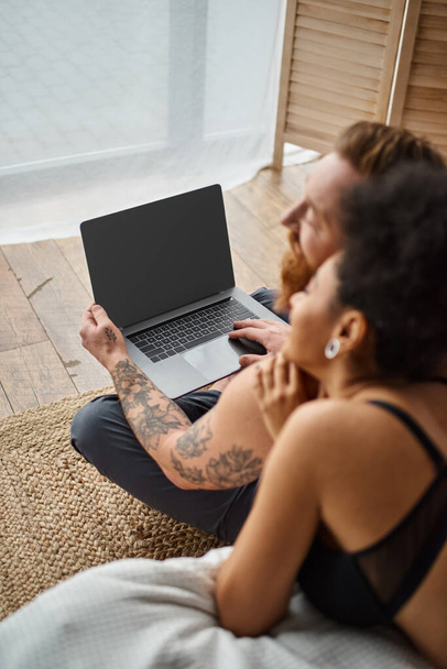 blurred interracial couple lying on bed and enjoying cozy moment while watching movie on laptop - Photo, Image