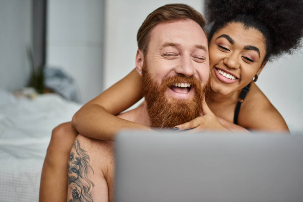 getatoeëerde man en Afrikaans Amerikaanse vrouw kijken comedy film op laptop en samen lachen - Foto, afbeelding