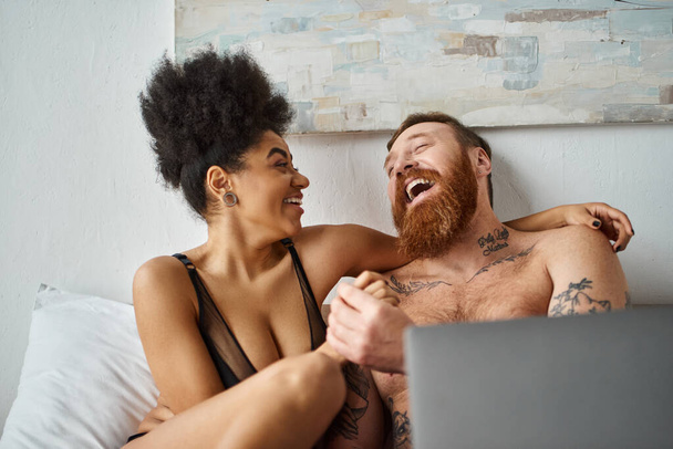 Χαρούμενη Αφροαμερικανή γυναίκα με εσώρουχα που χρησιμοποιεί φορητό υπολογιστή και ξαπλώνει στο κρεβάτι κοντά σε άντρα με τατουάζ, γελάει - Φωτογραφία, εικόνα