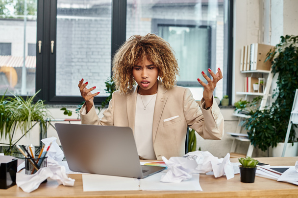 irritated curly african american businesswoman at her desk with laptop and crumpled papers, stress - Photo, Image