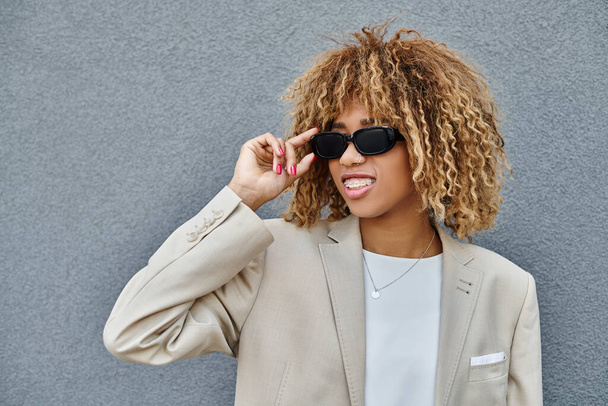 mujer afroamericana feliz con aparatos ortopédicos y pelo rizado con gafas de sol cerca del edificio de oficinas - Foto, Imagen