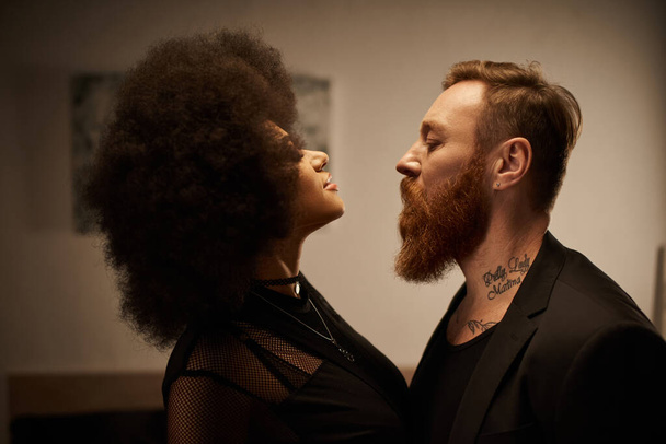 curly african american woman in chic dress seducing tattooed boyfriend with beard, date night - Photo, Image