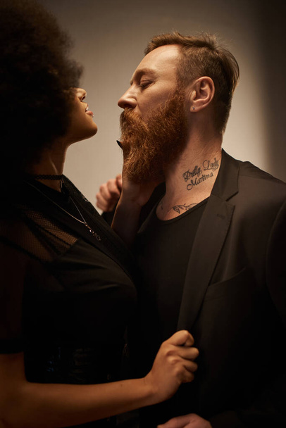 curly african american woman in black dress seducing tattooed boyfriend with beard, date night - Photo, Image
