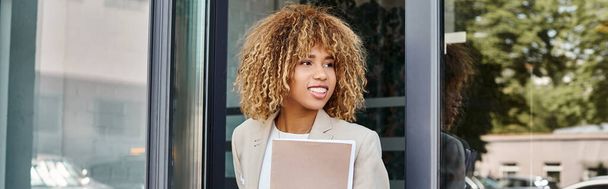 щаслива афроамериканська бізнес-леді з кучерявим волоссям, що тримає папку і виходить з будівлі, банер - Фото, зображення