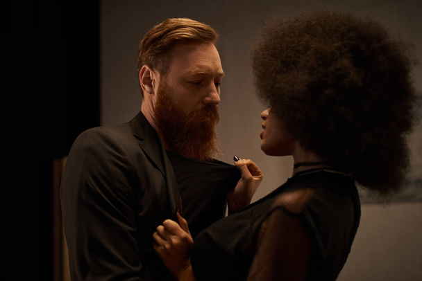 curly african american woman in dress pulling blazer of bearded man during date, sexy couple - Photo, Image
